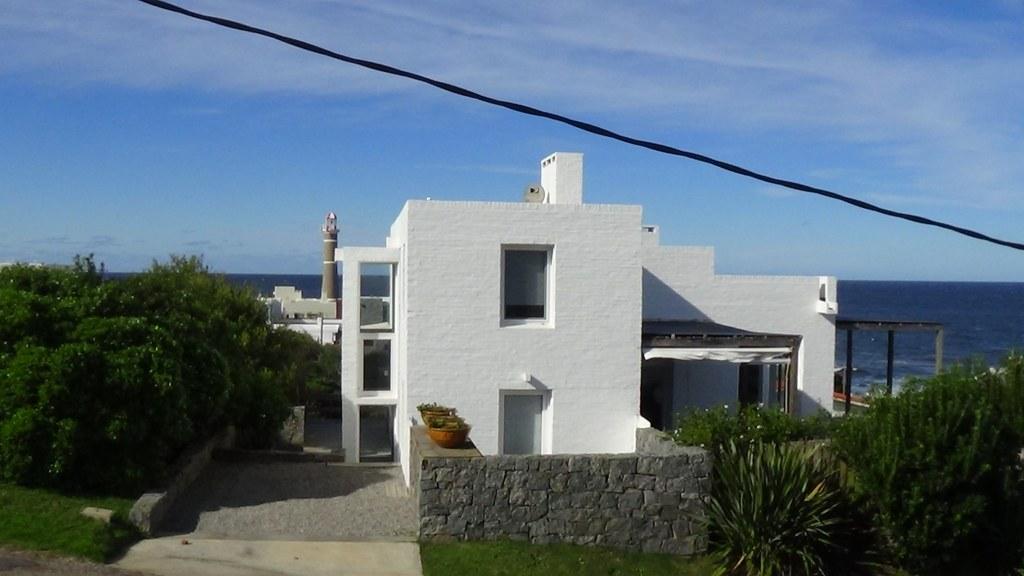 Casa blanca de ladrillo visto frente al mar en José Ignacio, con el faro al fondo y un muro de piedra en primer plano