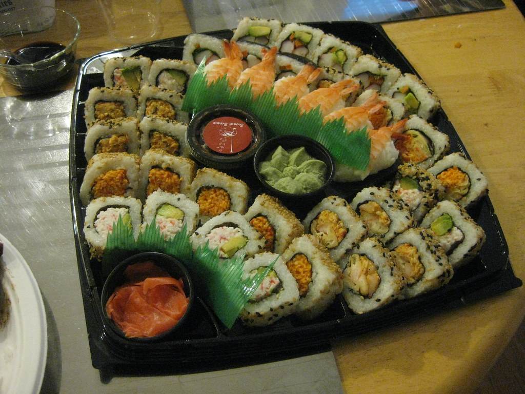 Bandeja negra con rolls de sushi con sésamo, nigiris de langostino, wasabi, jengibre encurtido y un pote de salsa sobre una mesa de madera