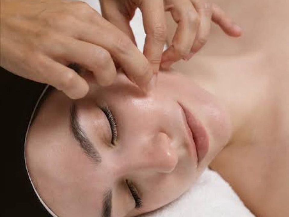 Hände massieren die Schläfen einer Frau mit geschlossenen Augen, die auf einem weißen Handtuch liegt, bei einer Kobido-Gesichtsmassage