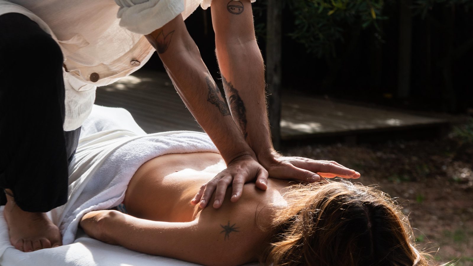 Guido D'Alessandro in weißem Hemd und mit tätowierten Armen legt die Hände auf den Rücken einer Frau, die im Freien auf einer Massageliege liegt, im Hintergrund Bäume